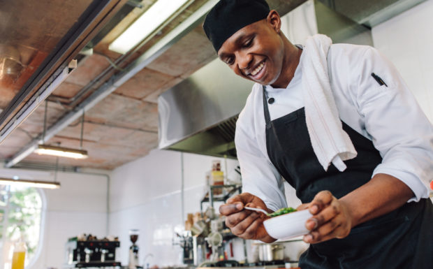 Gourmet chef in uniform cooking in a commercial kitchen. Happy male cook wearing apron standing by kitchen counter preparing food.